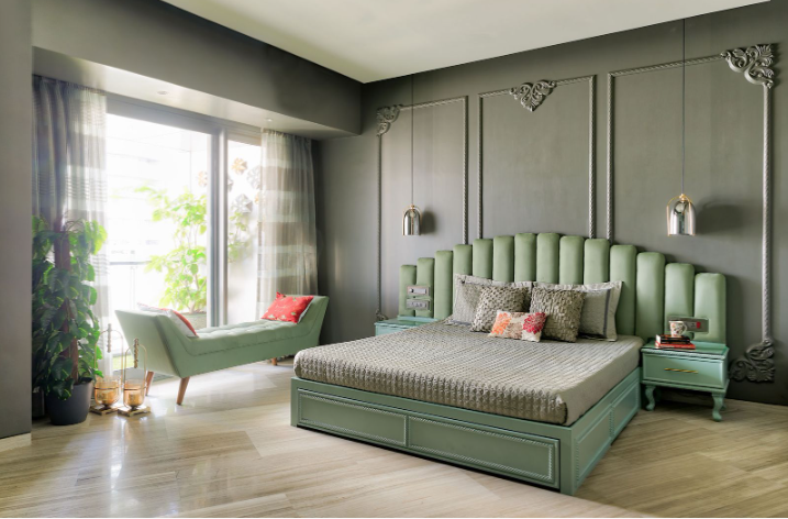 Spacious bedroom interior featuring an olive-green fluted headboard bed framed by dark gray decorative wall moldings, paired nightstands, and a light green chaise near a sunlit window.