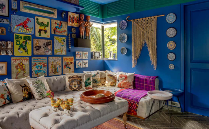 Cozy living room nook featuring vibrant blue walls adorned with retro superhero posters and macramé art, centered around a tufted cream L-shaped sofa styled with colorful patterned cushions.