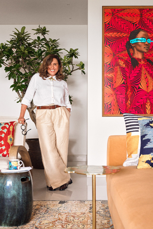 Portrait of interior designer Vinita Chaitanya dressed in a white shirt and wide-leg trousers, standing next to a potted ficus plant and an orange sofa with a vivid red pop art painting in the background.