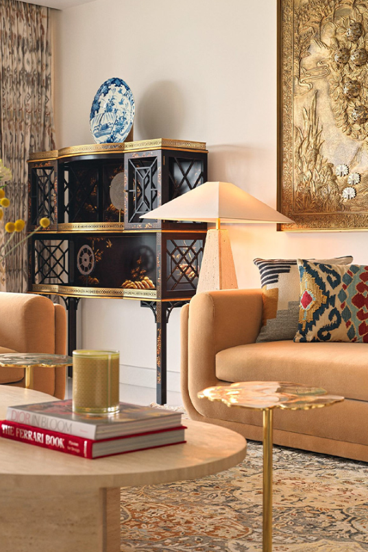 Living room interior showcasing an ornate black and gold curio cabinet topped with a blue-and-white porcelain plate, positioned behind a warm ochre sofa, a pyramid-shade floor lamp, and a travertine center table.