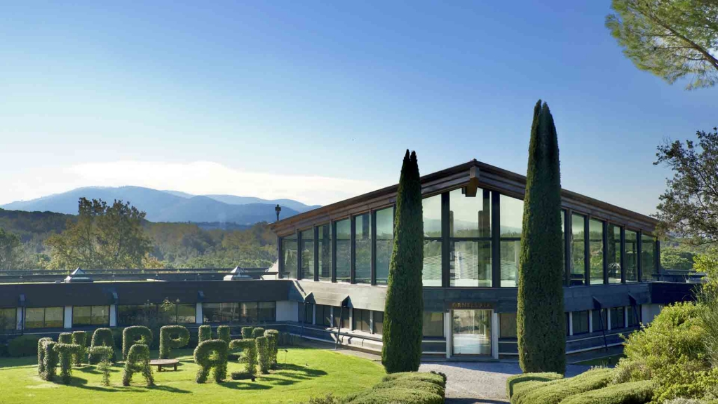 Modern glass winery building flanked by tall cypress trees with green lawns and distant blue mountains, prestigious wine estates