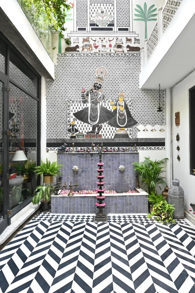 Bright open atrium with striking black and white chevron floor tiles, featuring an ornate wall mural depicting Lord Shrinathji above a mosaic water basin decorated with flower garlands.