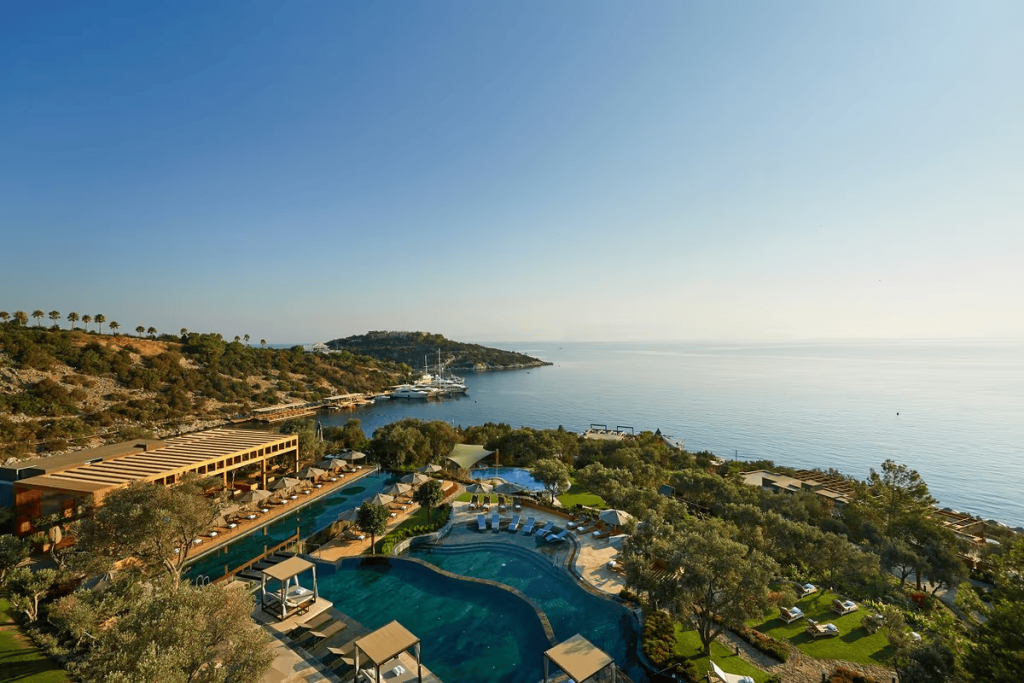 An elevated panoramic view of a hillside resort overlooking a turquoise bay dotted with yachts and islands, featuring multiple terraced swimming pools, sun decks with cabanas, and lush Mediterranean vegetation under a clear sky. Luxury hotel chains.