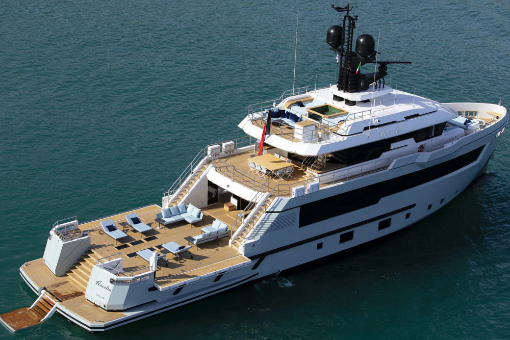 An aerial, rear three-quarter view of a large white explorer yacht. The yacht features an expansive teak aft deck with lounge seating and sunbeds, multiple staircases leading down to a swim platform, and an upper deck with a dining area. The name "AURORA" is visible on the stern. It is anchored in calm, blue-green water near a coastline. Cannes Yachting Festival.