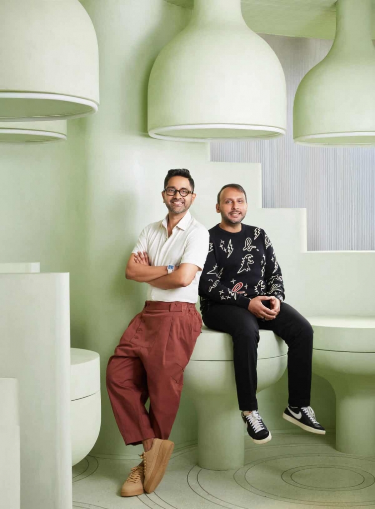 Two male designers posing together in a modern, light-green interior space with sculptural, bulbous seating and large pendant lamps. design democracy