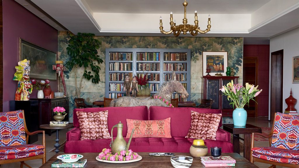 Opulent colorful living room with a rich magenta sofa adorned with patterned pillows, flanked by printed armchairs and styled with a wooden coffee table, background library shelving, and an ornate carved horse figure.