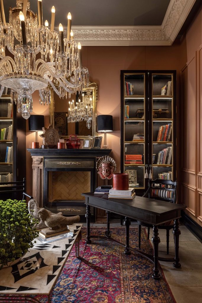 Elegant room interior showcasing a large crystal chandelier, a dark wooden desk over a colorful Persian area rug, a fireplace mantel with mirror, and tall illuminated glass-door bookcases.