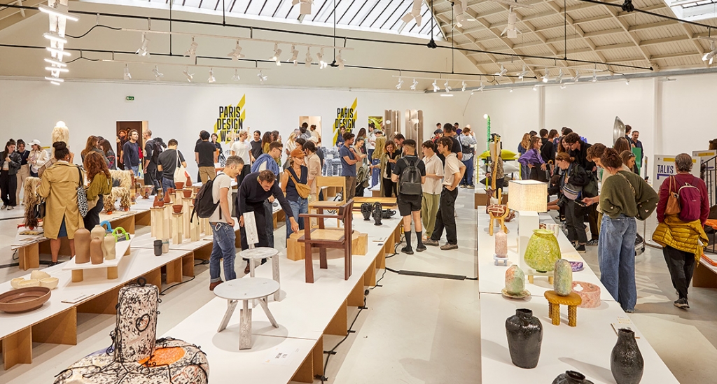 A crowded interior exhibition hall during an event, featuring long display tables filled with various ceramic vases, handmade chairs, and contemporary art objects. Visitors walk around browsing the exhibits under a large industrial skylight roof, with "Paris Design Week" signage visible on the back wall. Paris design week.