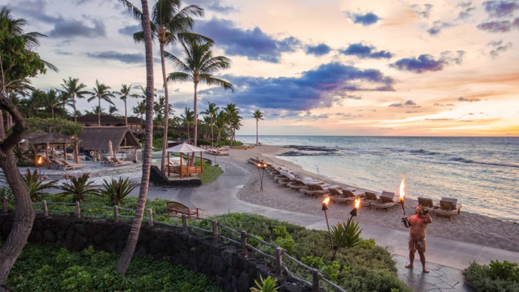 A Hawaiian beachfront resort at sunset, with a torch-lit path leading past thatched-roof huts, palm trees, and rows of lounge chairs, as a person in traditional attire performs with a fire torch, and the ocean reflects the colorful sky. Luxury hotel chains.