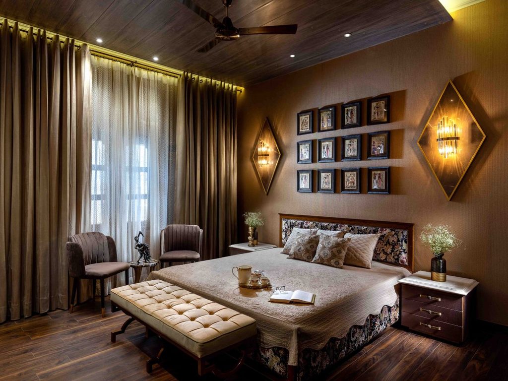 Master bedroom interior with dark wood flooring, taupe curtains, a tufted bench at the foot of the bed, and warm illuminated wall sconces flanking framed artwork.