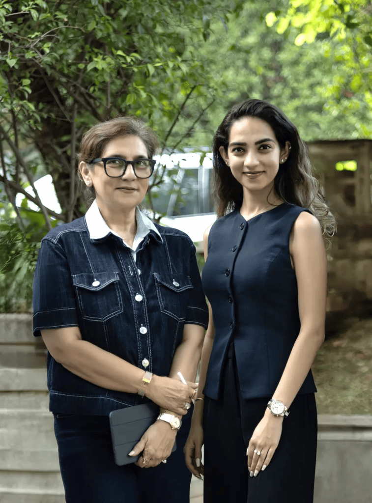 Two women standing side by side outdoors against a backdrop of lush green foliage; an older woman wearing glasses and a dark denim shirt, and a younger woman wearing a navy blue sleeveless top. design democracy