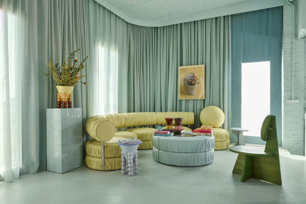 A modern living room interior styled in pastel tones, featuring a curved yellow sectional sofa, a light blue round coffee table, and soft mint-green curtains covering a curved wall. Paris design week.