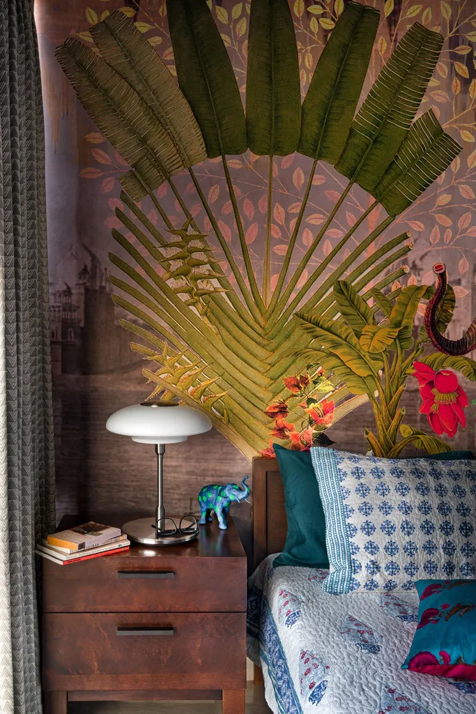 Bedside setup featuring an expressive tropical palm mural on the wall behind a bed dressed with blue patterned cushions, beside a two-drawer wooden nightstand topped with books and a mushroom-style table lamp.