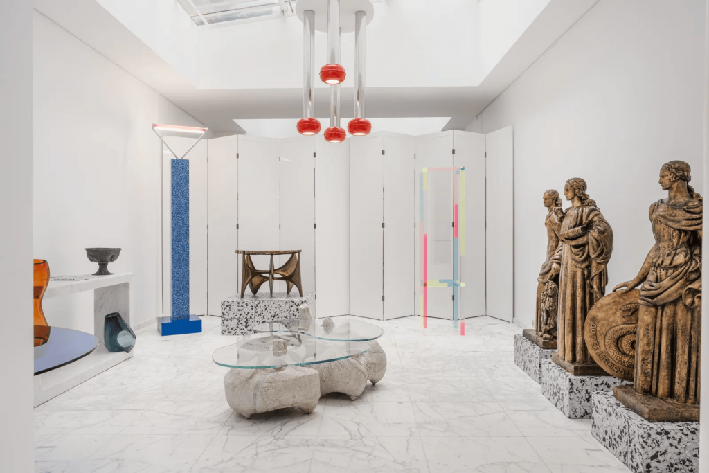 A bright contemporary art and design gallery interior with a white marble floor, featuring antique-style bronze statues, modern glass-topped stone coffee tables, a sculptural central pendant light fixture, and white folding screens. Paris design week.