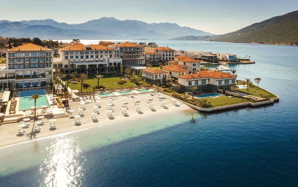 A sprawling coastal resort complex with cream-colored buildings and terracotta roofs, featuring multiple terraced swimming pools and a pristine white sand beach with umbrellas and loungers, overlooking a wide bay and distant mountains under a bright sun. Luxury hotel chains.