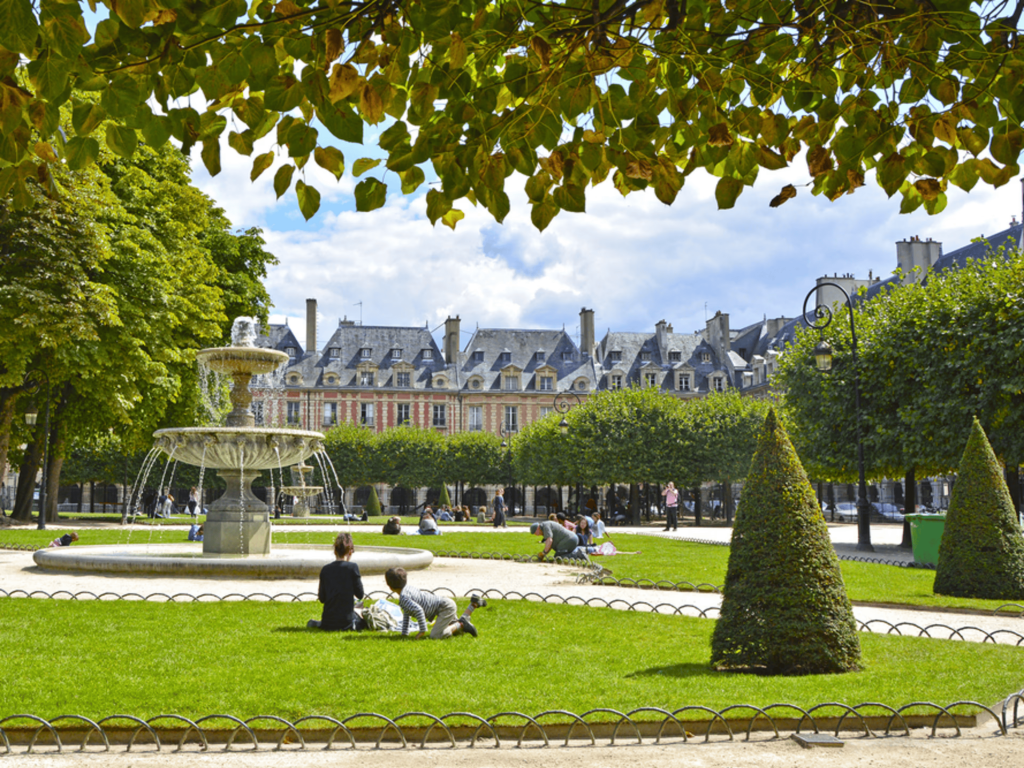 A sunny day at the Place des Vosges park in Paris, showing a central stone fountain, green lawns with people relaxing, neatly trimmed conical bushes, and historic red-brick residential buildings in the background under a cloudy sky. Paris design week.
