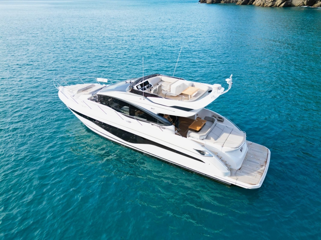 A high-angle, rear-quarter shot of a sleek white sports cruiser yacht. The yacht features a spacious cockpit with sunbeds, a helm station, and a large hydraulic swim platform. It is cruising at moderate speed through clear turquoise water, with a rocky coastline visible in the background. Cannes Yachting Festival.