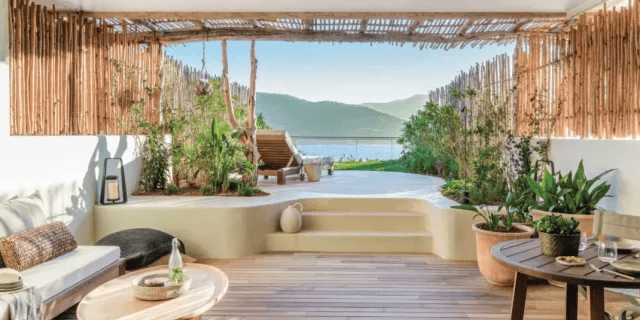 A luxurious, open-air hotel patio with light-colored stucco walls, wooden slat ceilings, and abundant potted plants, leading out to a private sun deck with a wooden lounge chair and a stunning sea view framed by a bamboo pergola. Luxury hotel chains.