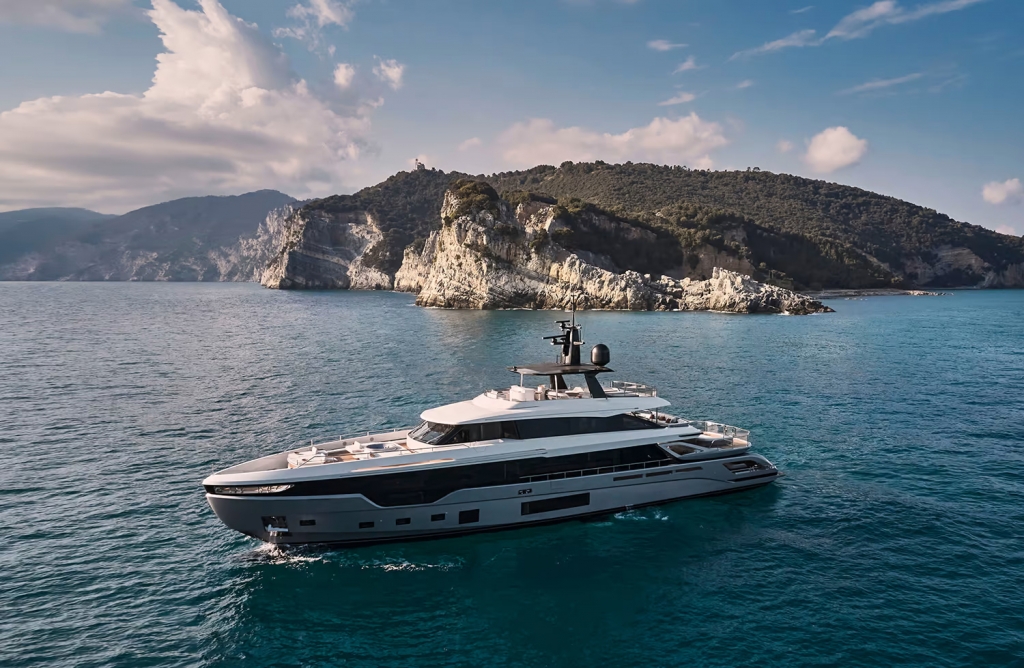 An aerial, three-quarter view of a large, modern superyacht with a white superstructure and dark hull, cruising near a scenic rocky coastline with green hills under a partly cloudy sky. monaco yacht show