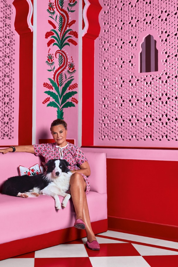 Designer Marie-Anne Oudejans sitting on a pink banquette with her black and white dog in a room styled with pink carved lattice screens, hand-painted floral motifs, and red checkered floors.