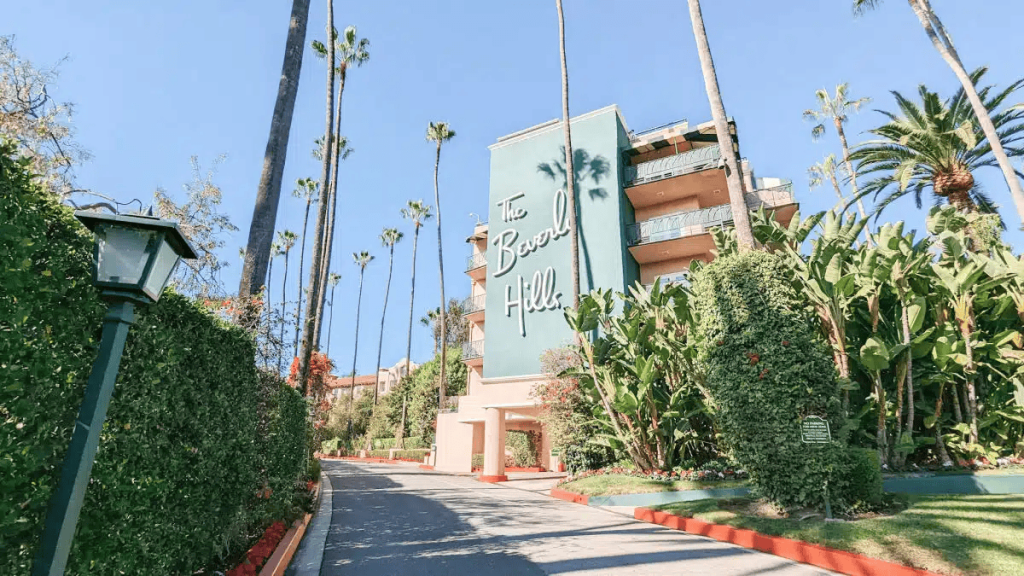 A lushly landscaped driveway curves toward the iconic pink facade of The Beverly Hills Hotel in California, flanked by tall palm trees and hedges under a clear blue sky. Luxury hotel chains.