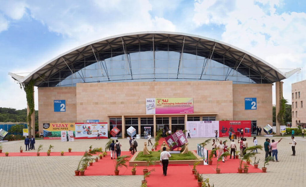 A wide shot of a modern convention center or exhibition hall with a large curved glass facade, a brick exterior, and a red carpet walkway decorated with potted plants and people walking outside. design democracy