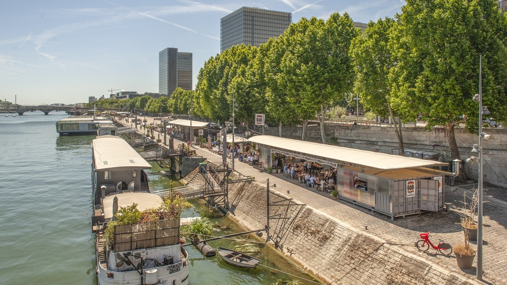 An urban waterfront view along the banks of the Seine river in Paris, featuring a modern pedestrian promenade, floating houseboats, outdoor riverside cafes, docked boats, and tall modern skyscrapers in the distance. Paris design week.
