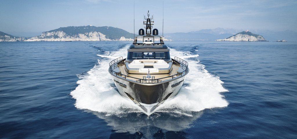 A modern, metallic-hulled superyacht cruises at speed across blue water, viewed head-on, creating a large white wake, with distant rocky islands under a clear sky. monaco yacht show