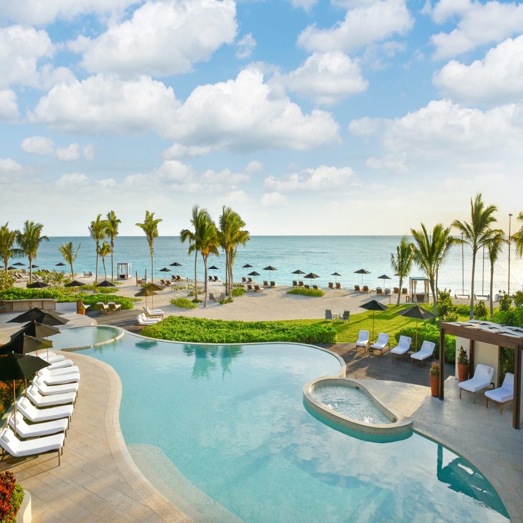 A luxurious, modern beachfront resort with a lagoon pool, white sand beach, and numerous lounge chairs, all framed by tall palm trees and turquoise ocean waters under a bright, cloudy sky. Luxury hotel chains.