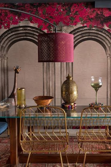 Dining area with two gold wire dining chairs in front of a glass-top table styled with an ornate brass egg-shaped urn and glassware, set against an archway wall mural and lit by a perforated pink drum light.