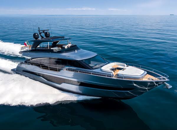 A high-angle, bow-on shot of a grey metallic motor yacht planing at high speed across turquoise water, creating a large white wake. The yacht features modern design, a sundeck with loungers, and radar domes. An Italian flag is visible on the stern. Cannes Yachting Festival.
