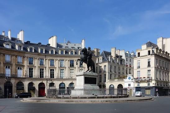 A sunny daytime street view of the Place Vendôme in Paris, featuring the iconic central bronze column and statue surrounded by historic Parisian Haussmann-style architecture under a clear blue sky. Paris design week.