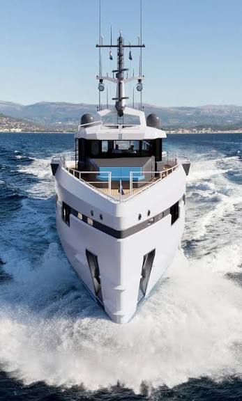 A close-up, head-on view of the bow of a modern white superyacht with a vertical bow design, cruising at speed and generating a large bow wake. monaco yacht show