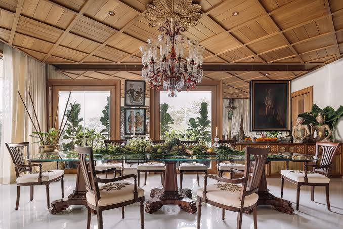 Stately dining room with a geometric coffered wood ceiling and large crystal chandelier hanging over a long glass-top pedestal table with lush botanical centerpieces and cream-cushioned armchairs.
