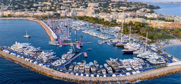 An aerial view of a luxurious sailing yacht gliding through the sea, showcasing its wooden deck and large branded sails at the Cannes Yachting Festival 2026.