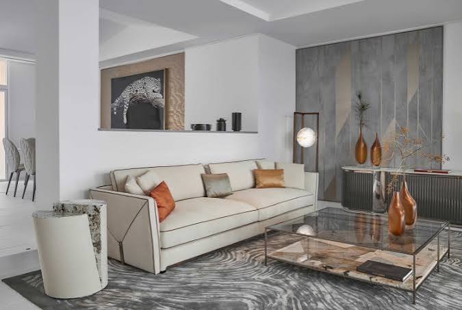 A contemporary living room featuring a cream sectional sofa with rust-colored pillows, a glass-top coffee table with a wooden base, and a patterned grey area rug at luxury furniture showrooms in Monaco.