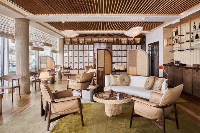 A warm, sophisticated lounge area with wooden slat ceilings, plush leather armchairs, a central woven coffee table, and shelving filled with decor at luxury furniture showrooms in Monaco.
