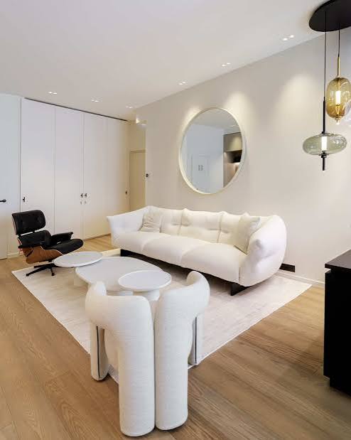 A minimalist living area with a white curved sofa, modern accent chairs, a round white coffee table, and a large circular wall mirror at luxury furniture showrooms in Monaco.