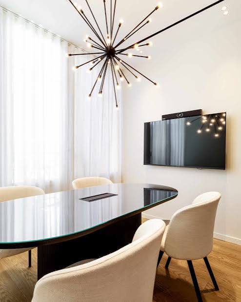 A sleek meeting room featuring an oval glass-top conference table surrounded by cream chairs beneath a striking modern sputnik chandelier, found in luxury furniture showrooms in Monaco.