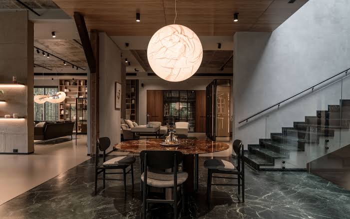 A sophisticated modern dining and living space featuring a dark marble dining table, black chairs, a large glowing spherical pendant lamp, and a marble staircase at luxury furniture showrooms in Hyderabad.
