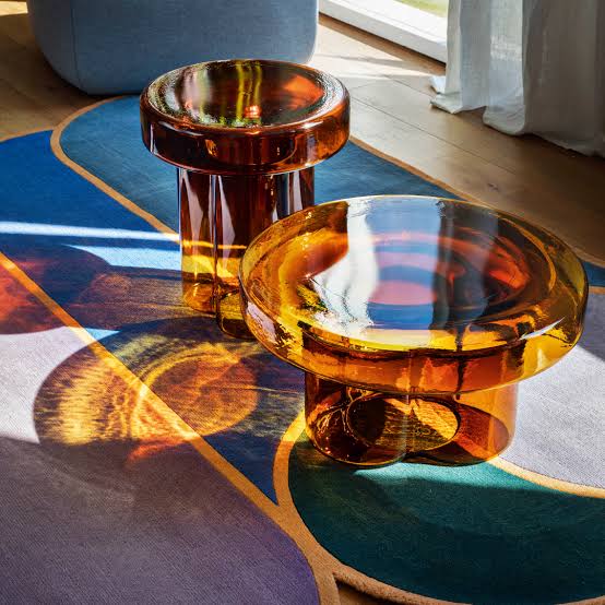 Two glossy amber glass side tables with mushroom-shaped tops resting on a vibrant blue and multi-colored area rug at luxury furniture showrooms in Hyderabad.