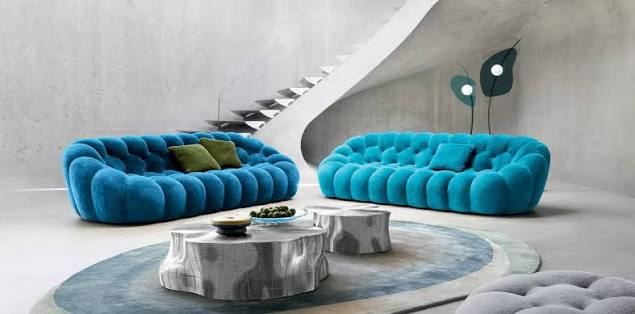 A spacious modern living room featuring two bright teal tufted bubble sofas, a unique tree-trunk style coffee table, and an abstract staircase in the background at luxury furniture showrooms in Hyderabad.