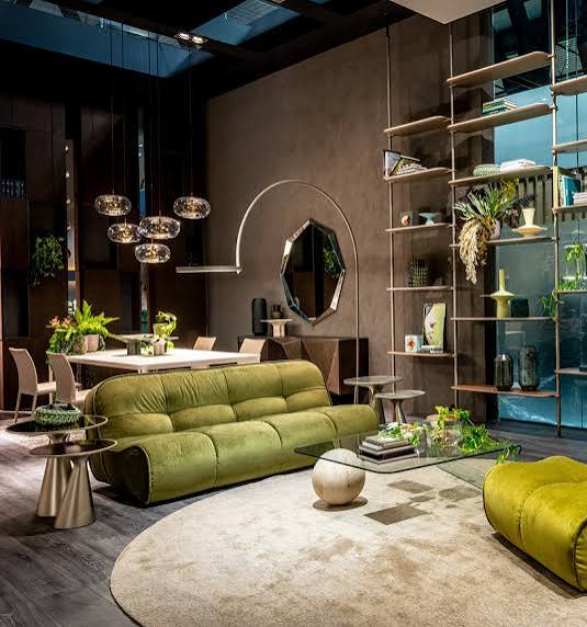 A modern lounge area showcasing a green velvet sofa, a large circular cream area rug, a minimalist glass coffee table, and an open metal bookshelf unit at luxury furniture showrooms in Hyderabad.