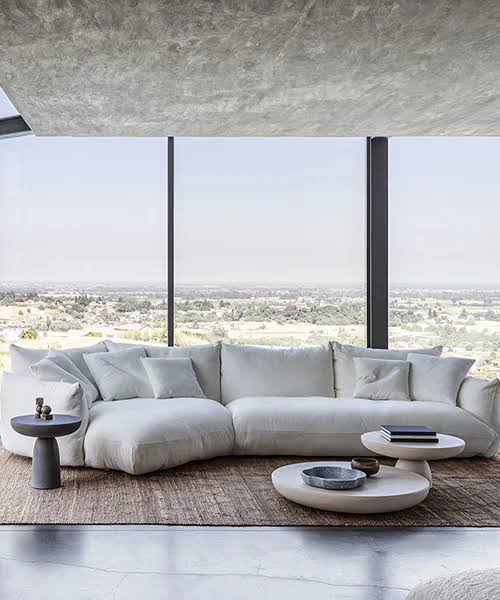 A minimalist living room with a large white modular sofa, contemporary round coffee tables, and floor-to-ceiling windows offering a sweeping panoramic view at luxury furniture showrooms in Hyderabad.
