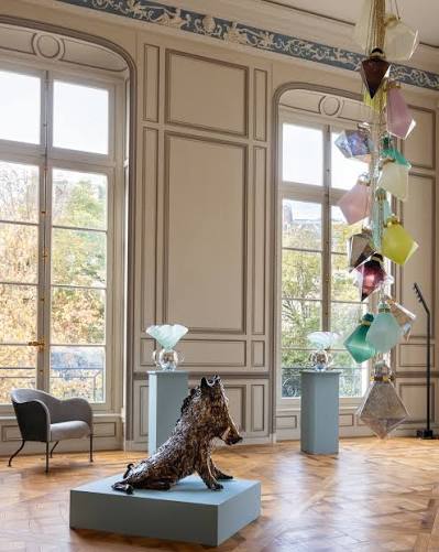 An elegant room with tall windows, high ceilings with molding, a hanging multicolored geometric glass mobile, and a dark sculpted animal figure on a low pedestal at paris design week