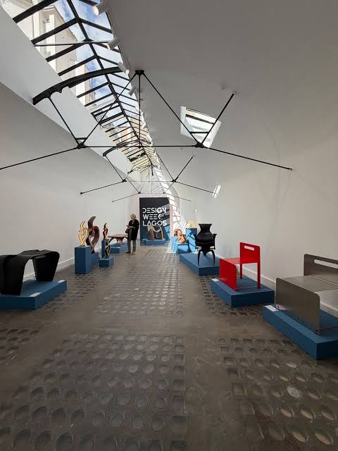 A minimalist exhibition space showcasing sleek, arched metallic and wooden objects on pedestals alongside a glowing illuminated arch, with a person walking past in the background at paris design week