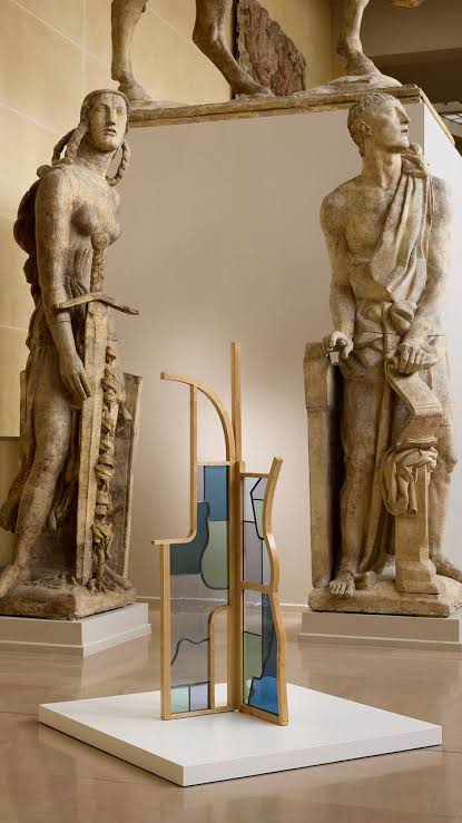 A modern wooden screen with glass inserts displayed on a white platform inside a classical museum gallery featuring antique stone statues in the background at paris design week