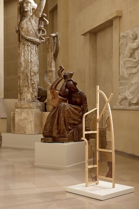 A contemporary multi-colored glass and wood screen displayed on a white pedestal, positioned between two classical stone statues of figures holding weapons at paris design week
