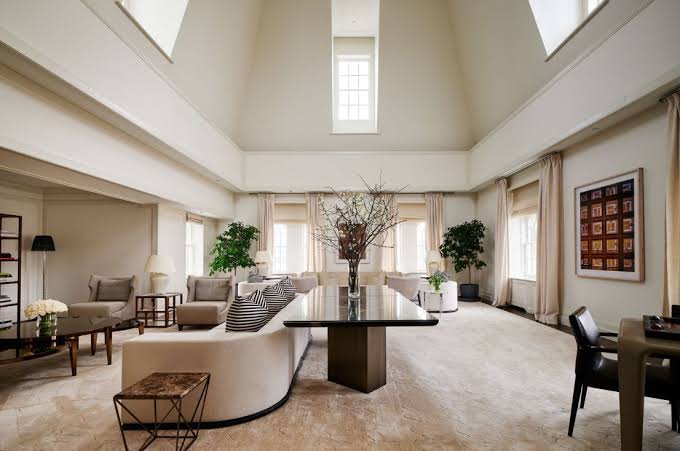 A sprawling, high-ceilinged penthouse living and dining room featuring white walls, a large curved sofa, a sleek long dining table, potted plants, and tall windows letting in natural light, all grouped under most prestigious hotel suites