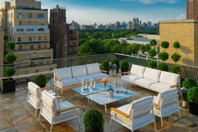 A rooftop terrace patio furnished with modern white outdoor sofas, armchairs, and coffee tables, surrounded by potted plants and overlooking a cityscape with green trees and buildings, all grouped under most prestigious hotel suites